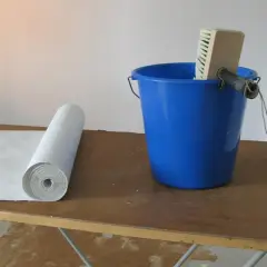 Vliestapete liegt bereit, daneben ein blauer Eimer mit Kleister und einer Bürste. Perfekt für die Renovierung von Raufasertapete.