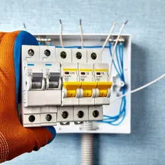 Sicherheitschalter werden in eine Verteilerdose eingebaut. Ein Handwerker mit orangefarbenem Handschuh hält die Sicherungen.