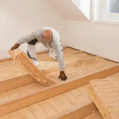 Mann verlegt Dämmmaterial auf einer Holzbalkendecke. Darauf kommt später der Trockenestrich.