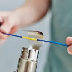 Hände erhitzen mit einem Heißluftgebläse einen gelben Schrumpfschlauch auf einem blauen Kabel.
