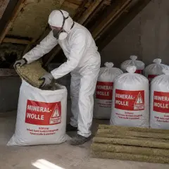 Mann in Schutzkleidung packt Mineralwolle in Säcke. Mehrere Säcke mit der Aufschrift "MINERALWOLLE" stehen bereit zur fachgerechten Entsorgung.