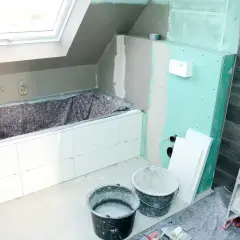 Badezimmer im Umbau: weiße Fliesen auf Rigips-Wänden, Badewanne abgedeckt, Eimer und Leiter bereit für die Fertigstellung.
