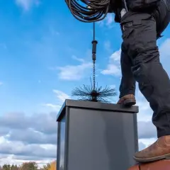 Mann auf dem Dach mit Schornsteinfegerbürste, bereit, den Schornstein selber zu reinigen.