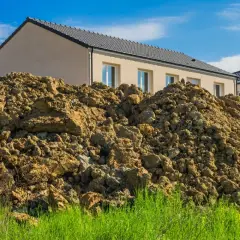Große Haufen erdreich aus einer Baugrube türmen sich vor neuen Häusern unter blauem Himmel.