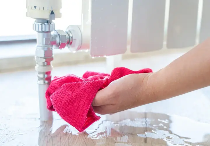 Hand wischt Wasser auf dem Boden auf, das aus einem Heizungsrohr tropft. Vorsicht, wann frieren Heizungsrohre ein!