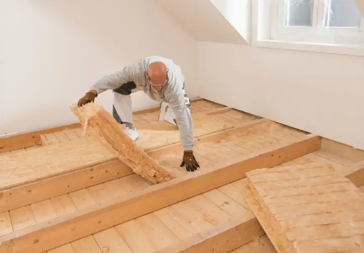 Mann verlegt Dämmmaterial auf einer Holzbalkendecke. Darauf kommt später der Trockenestrich.