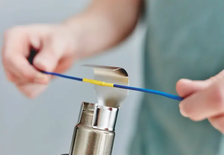 Hände erhitzen mit einem Heißluftgebläse einen gelben Schrumpfschlauch auf einem blauen Kabel.