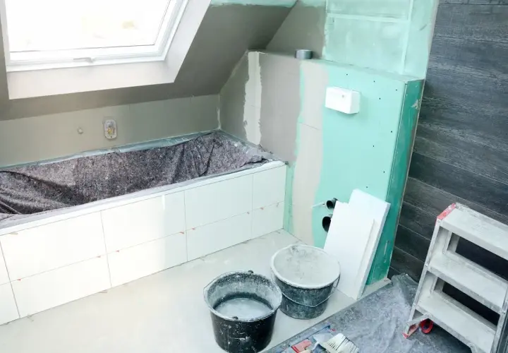 Badezimmer im Umbau: weiße Fliesen auf Rigips-Wänden, Badewanne abgedeckt, Eimer und Leiter bereit für die Fertigstellung.