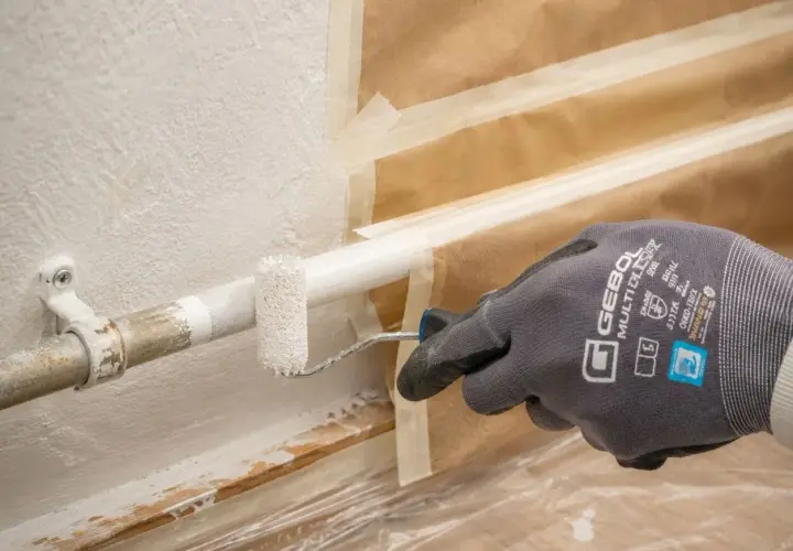 Mit einem kleinen Roller werden Heizungsrohre weiß gestrichen. Die Wand ist abgeklebt, um sie vor Farbspritzern zu schützen.