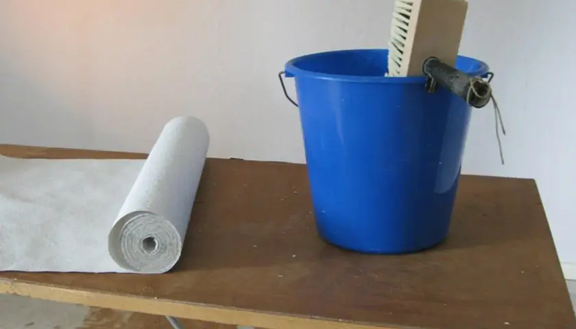 Vliestapete liegt bereit, daneben ein blauer Eimer mit Kleister und einer Bürste. Perfekt für die Renovierung von Raufasertapete.