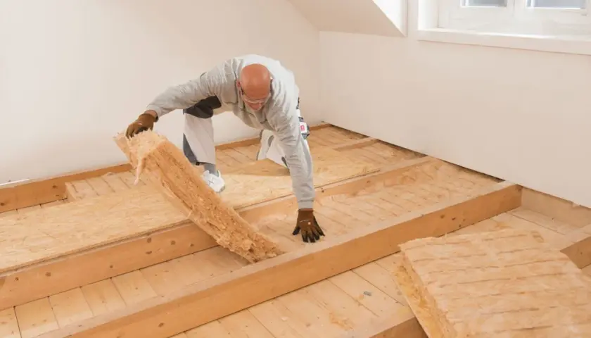 Mann verlegt Dämmmaterial auf einer Holzbalkendecke. Darauf kommt später der Trockenestrich.