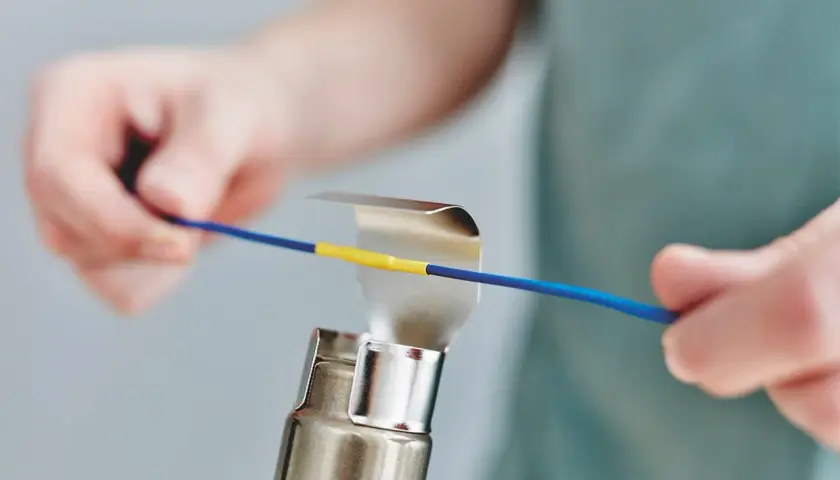 Hände erhitzen mit einem Heißluftgebläse einen gelben Schrumpfschlauch auf einem blauen Kabel.