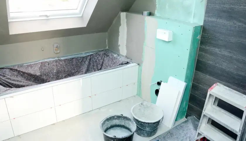 Badezimmer im Umbau: weiße Fliesen auf Rigips-Wänden, Badewanne abgedeckt, Eimer und Leiter bereit für die Fertigstellung.
