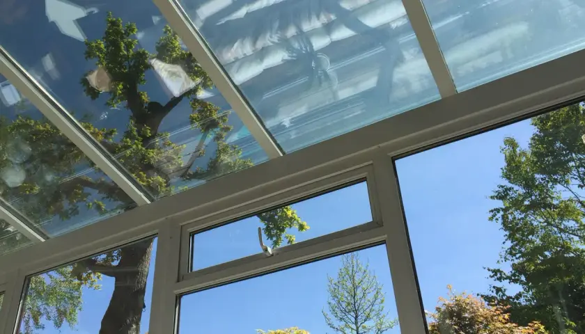 Blick durch ein Fenster in einen Wintergarten mit Glasdach. Bäume spiegeln sich im Glas. Dichtungsarbeiten am Wintergarten sind wichtig.