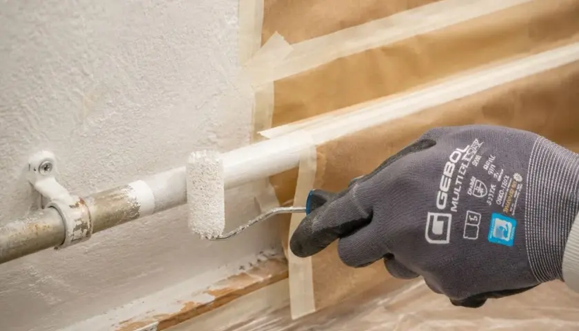 Mit einem kleinen Roller werden Heizungsrohre weiß gestrichen. Die Wand ist abgeklebt, um sie vor Farbspritzern zu schützen.