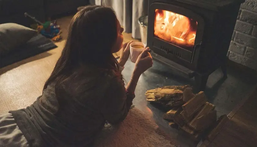 Gemütliches Beisammensein am Kaminfeuer. Eine Frau genießt eine Tasse Tee, während sie die Wärme des Feuers spürt. Heizen ohne Strom ist eine romantische Alternative.