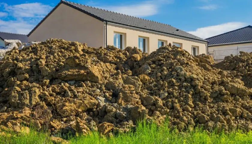 Große Haufen erdreich aus einer Baugrube türmen sich vor neuen Häusern unter blauem Himmel.