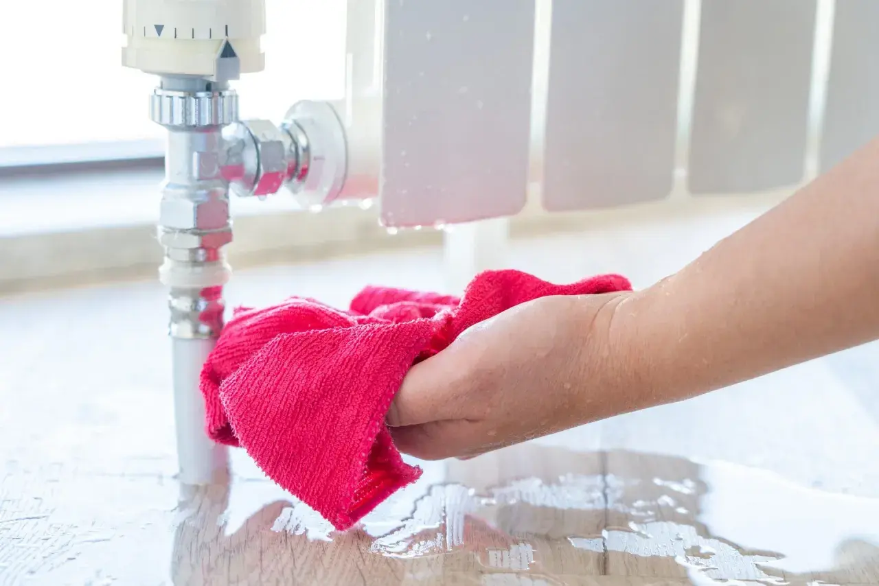 Hand wischt Wasser auf dem Boden auf, das aus einem Heizungsrohr tropft. Vorsicht, wann frieren Heizungsrohre ein!