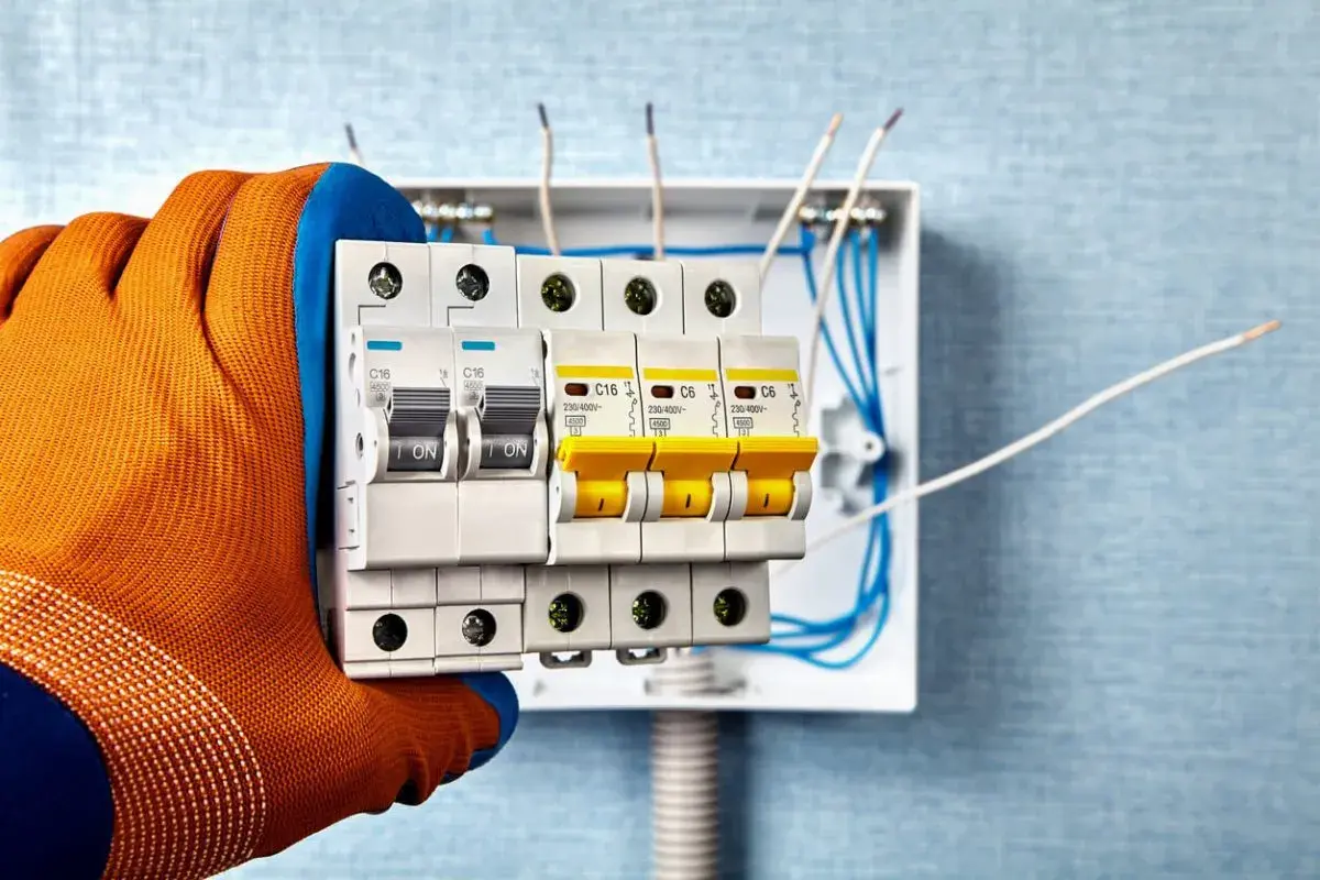 Sicherheitschalter werden in eine Verteilerdose eingebaut. Ein Handwerker mit orangefarbenem Handschuh hält die Sicherungen.