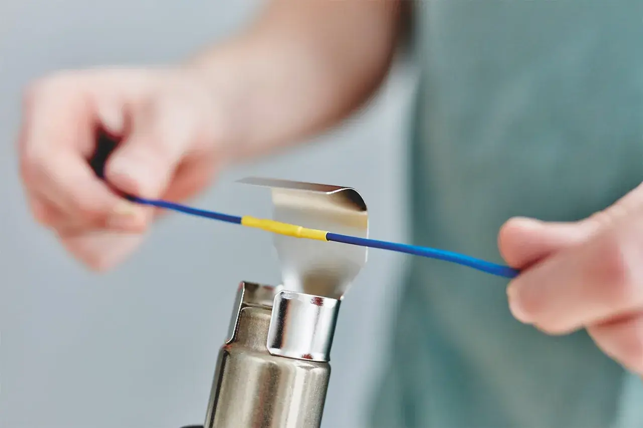 Hände erhitzen mit einem Heißluftgebläse einen gelben Schrumpfschlauch auf einem blauen Kabel.