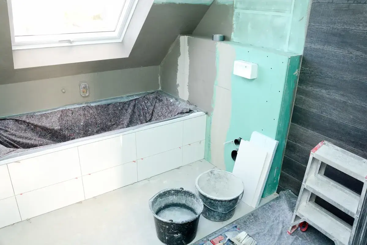 Badezimmer im Umbau: weiße Fliesen auf Rigips-Wänden, Badewanne abgedeckt, Eimer und Leiter bereit für die Fertigstellung.