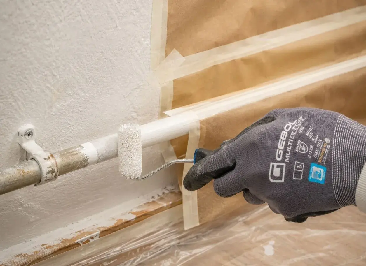 Mit einem kleinen Roller werden Heizungsrohre weiß gestrichen. Die Wand ist abgeklebt, um sie vor Farbspritzern zu schützen.