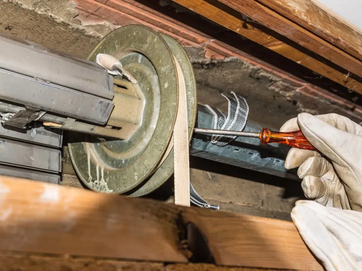 Hand mit Handschuh schraubt am Rollladen. Der alte Rolladengurt wird gewechselt, um den Rollladen wieder funktionsfähig zu machen.