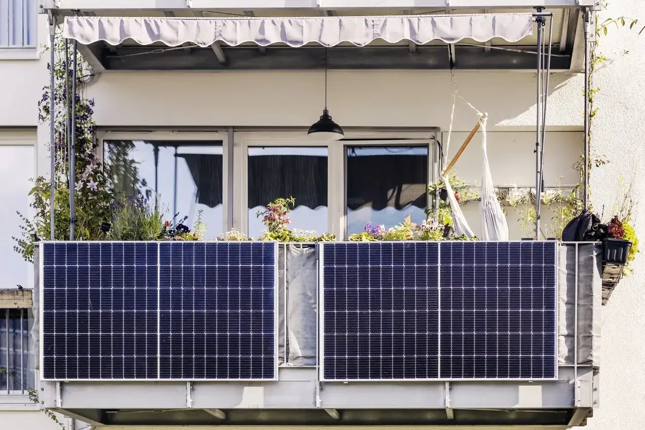 Zwei Solarmodule sind am Balkongel&auml;nder befestigt. Davor bl&uuml;hen Blumen und Pflanzen. Ein H&auml;ngesessel und eine Lampe vervollst&auml;ndigen das Bild.