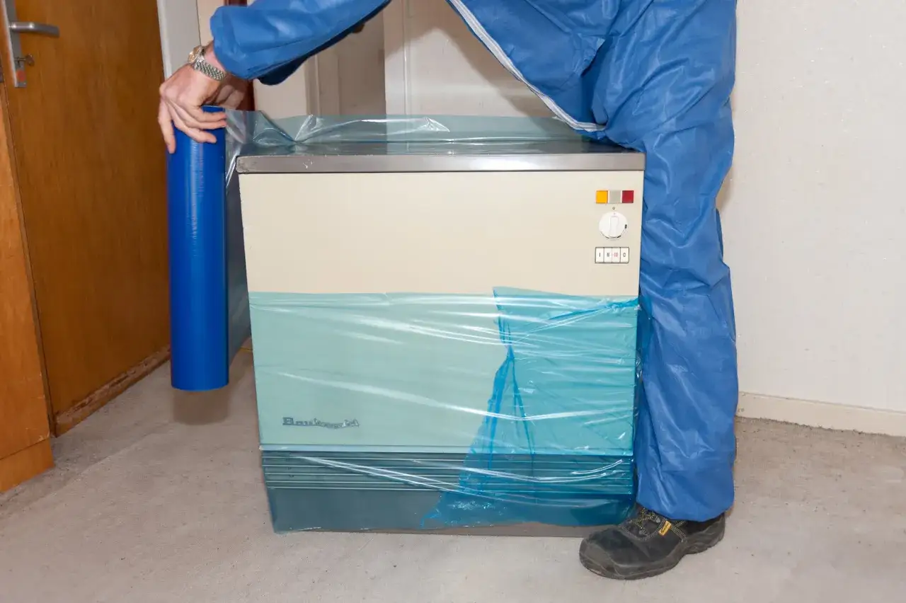 Mann in blauer Schutzkleidung wickelt einen alten Nachtspeicherofen in Folie ein, um ihn fachgerecht zu entsorgen.
