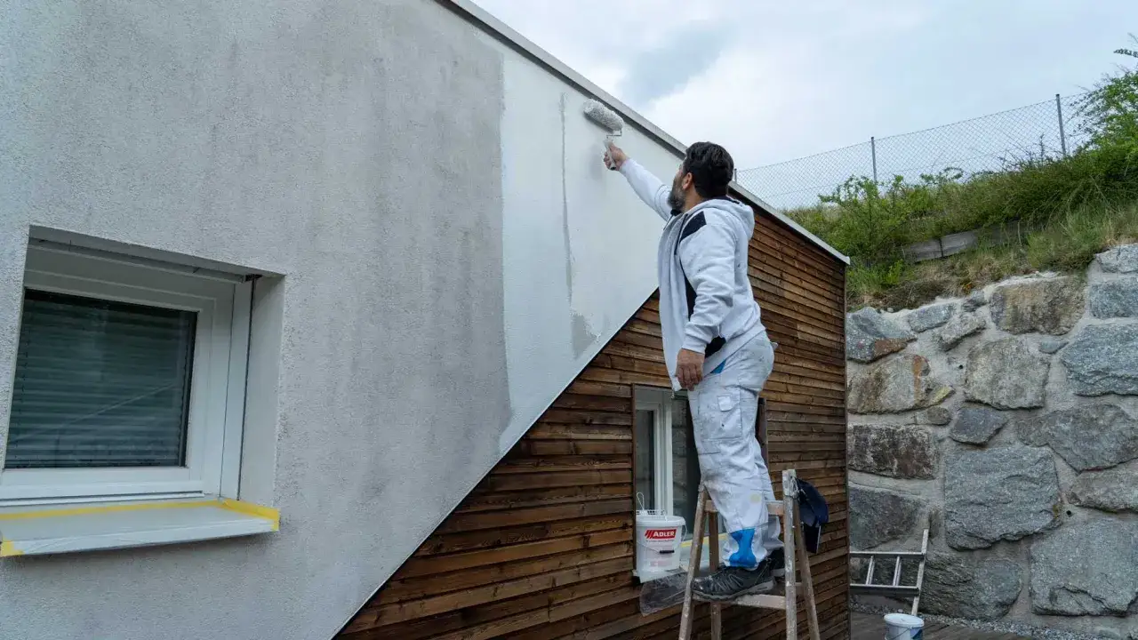 Mann streicht Fassade mit Grundierung. Ein Eimer Farbe steht bereit.